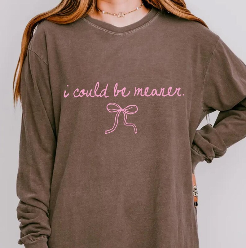 May include: A brown long-sleeve shirt with the text "i could be meaner" in pink script, along with a pink bow graphic. The shirt has a crew neck and long sleeves.