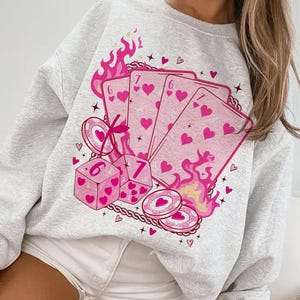 May include: Light gray sweatshirt featuring a pink graphic of playing cards, dice, and casino chips. The cards display hearts and the numbers six and seven. Pink flames and hearts surround the design.