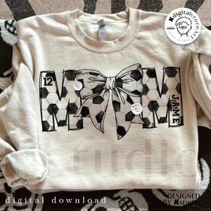 Peut inclure: Un design noir et blanc du mot "MOM" avec un motif de ballon de football. Le "M" porte le num&eacute;ro "12" et le "M" porte le nom "JAMIE". Un n&oelig;ud noir et blanc &agrave; motif de ballon de football se trouve au centre du design.
