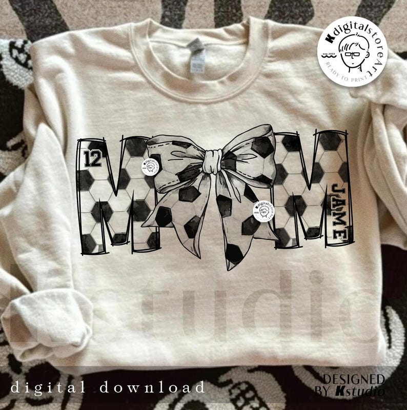 Peut inclure: Un design noir et blanc du mot "MOM" avec un motif de ballon de football. Le "M" porte le num&eacute;ro "12" et le "M" porte le nom "JAMIE". Un n&oelig;ud noir et blanc &agrave; motif de ballon de football se trouve au centre du design.