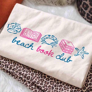 May include: Cream-colored tote bag with a blue and pink graphic that reads "beach book club." The design includes illustrations of a flower, open book, crab, stack of books, and starfish. The bag is folded on top of a leopard print fabric.