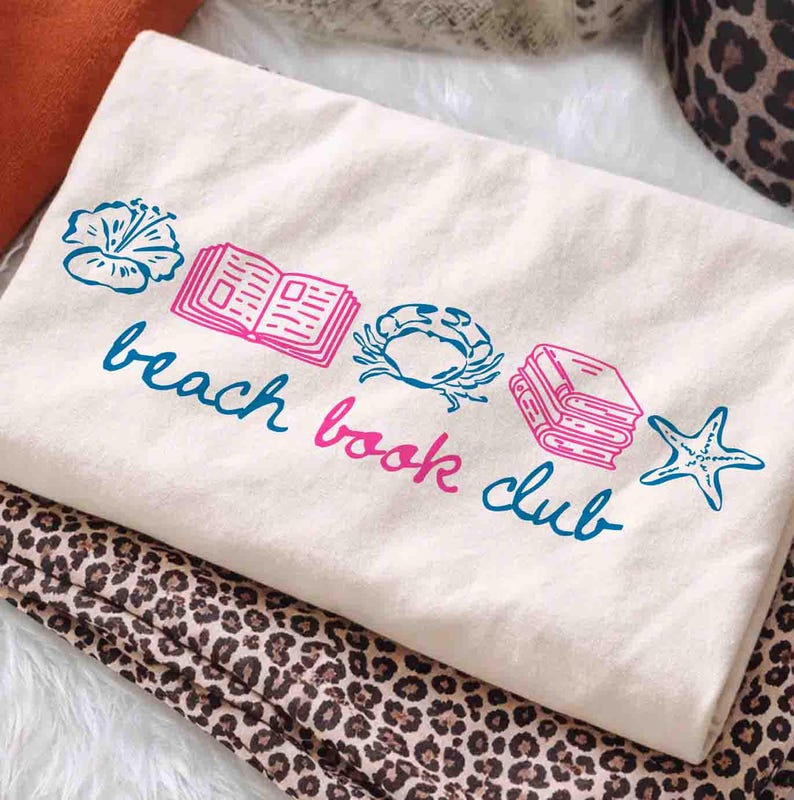 May include: Cream-colored tote bag with a blue and pink graphic that reads "beach book club." The design includes illustrations of a flower, open book, crab, stack of books, and starfish. The bag is folded on top of a leopard print fabric.