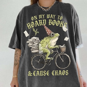 May include: Dark grey t-shirt with the slogan "ON MY WAY TO HOARD BOOKS & CAUSE CHAOS." The graphic shows a green frog on a bicycle, carrying books and wearing a wizard's hat. A small dragon and floating papers complete the design.