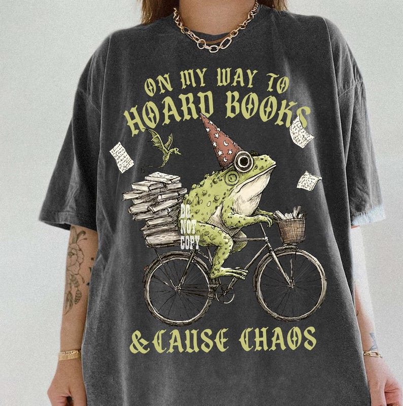 May include: Dark grey t-shirt with the slogan "ON MY WAY TO HOARD BOOKS & CAUSE CHAOS." The graphic shows a green frog on a bicycle, carrying books and wearing a wizard's hat. A small dragon and floating papers complete the design.