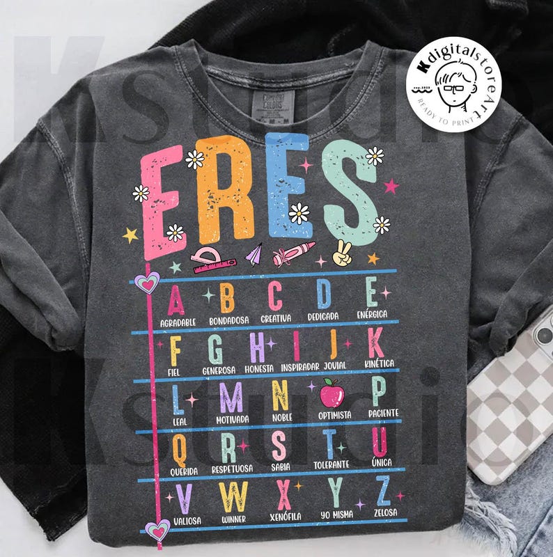 May include: Dark gray sweatshirt with the word "ERES" in colorful letters, decorated with flowers and school supplies. Below, an alphabet with positive adjectives in Spanish. A casual, comfortable garment.