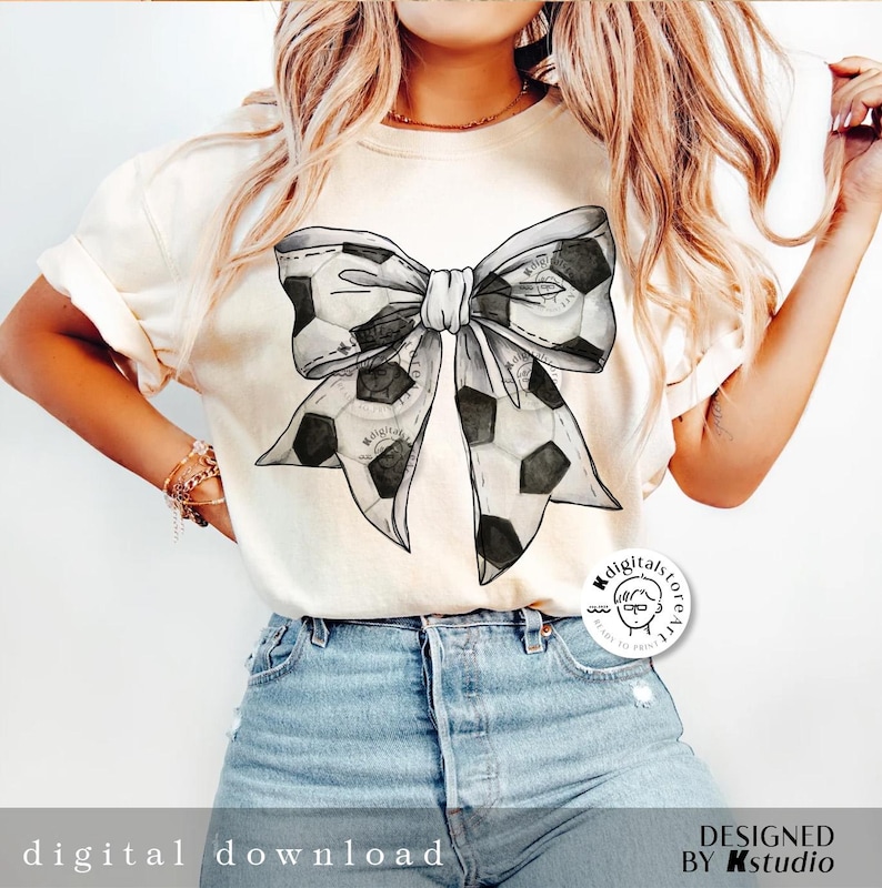 May include: A white t-shirt with a black and white soccer ball bow design. The shirt has a crew neck and short sleeves. The design is centered on the shirt. The shirt is being worn by a person with long blonde hair. The shirt has a small logo on the bottom right corner that says "Kdigitalstore" with a cartoon person wearing glasses. The text "DESIGNED BY Kstudio" is at the bottom of the image.
