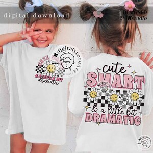 May include: White t-shirt with a pink and black design featuring the text "cute SMART & a little bit DRAMATIC". The design includes a checkered pattern, daisies with smiley faces, and pink stars.