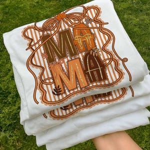 Op de afbeelding: Een stapel witte t-shirts met een herfstthema-afbeelding. Het ontwerp toont het woord "MAMA" in bruine en oranje letters, versierd met linten, bladeren en een gestreepte achtergrond. De shirts zijn netjes opgevouwen.