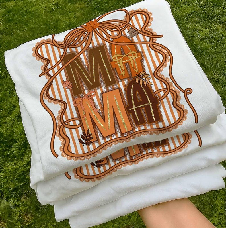 Op de afbeelding: Een stapel witte t-shirts met een herfstthema-afbeelding. Het ontwerp toont het woord "MAMA" in bruine en oranje letters, versierd met linten, bladeren en een gestreepte achtergrond. De shirts zijn netjes opgevouwen.