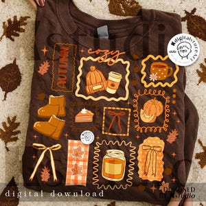 May include: A brown sweatshirt with autumn-themed illustrations. The design includes images of a hat, coffee cup, boots, pumpkin, pie, and leaves. Text includes "Autumn", "cozy", and "digital download".