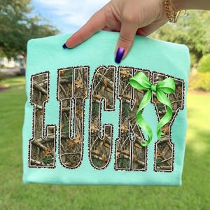 May include: A light teal sweatshirt with the word "LUCKY" in a camouflage pattern with a leopard print border. A green bow is on the right side of the word. The sweatshirt is held up by a hand.