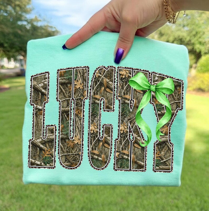 May include: A light teal sweatshirt with the word "LUCKY" in a camouflage pattern with a leopard print border. A green bow is on the right side of the word. The sweatshirt is held up by a hand.