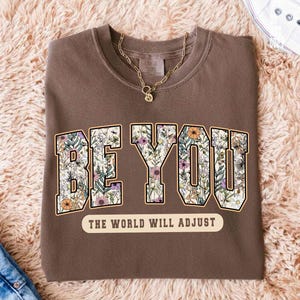 May include: A brown sweatshirt with the words "BE YOU" in a floral design. Below the text is the phrase "THE WORLD WILL ADJUST" in a cream-colored banner. A gold necklace is draped on the shirt.