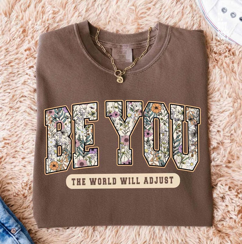 May include: A brown sweatshirt with the words "BE YOU" in a floral design. Below the text is the phrase "THE WORLD WILL ADJUST" in a cream-colored banner. A gold necklace is draped on the shirt.