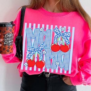 May include: A bright pink sweatshirt featuring the word "MOM" in a blue gingham pattern with red cherries and blue bows. White flowers and vertical stripes accent the design. The sweatshirt is a vibrant, eye-catching piece.