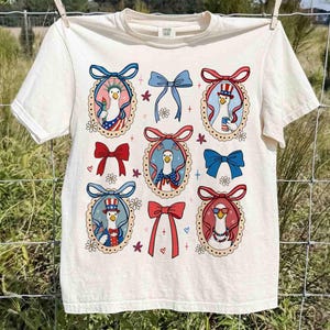 Op de afbeelding: Cr&egrave;mekleurig T-shirt met een patriottisch ganzenontwerp. Het shirt toont meerdere ovale kaders met ganzenillustraties in patriottische outfits, omringd door strikken en bloemmotieven. Het ontwerp gebruikt de kleuren rood, wit en blauw.