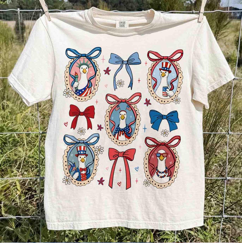 Op de afbeelding: Cr&egrave;mekleurig T-shirt met een patriottisch ganzenontwerp. Het shirt toont meerdere ovale kaders met ganzenillustraties in patriottische outfits, omringd door strikken en bloemmotieven. Het ontwerp gebruikt de kleuren rood, wit en blauw.