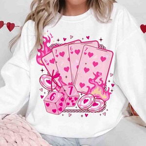 May include: White sweatshirt featuring a pink graphic of playing cards, dice, and poker chips with heart symbols. Pink flames and decorative elements surround the design. The playing cards show the numbers 6 and 7.