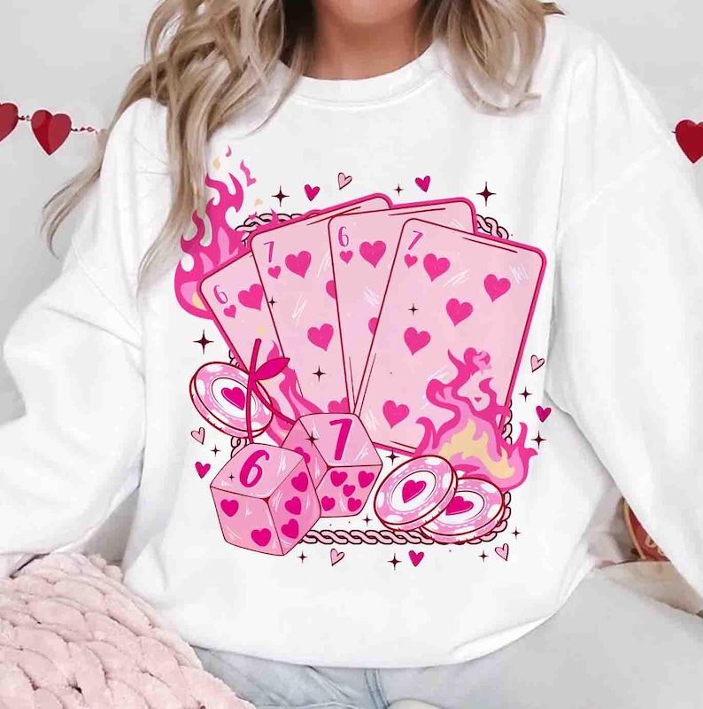 May include: White sweatshirt featuring a pink graphic of playing cards, dice, and poker chips with heart symbols. Pink flames and decorative elements surround the design. The playing cards show the numbers 6 and 7.