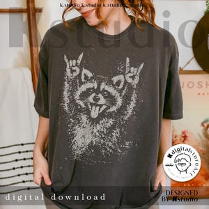 Pu&ograve; includere: T-shirt grigio scuro con una grafica usurata di un procione che fa gesti rock and roll. Il procione ha la lingua di fuori. Le parole "digital download" sono in basso. Design di Kstudio.
