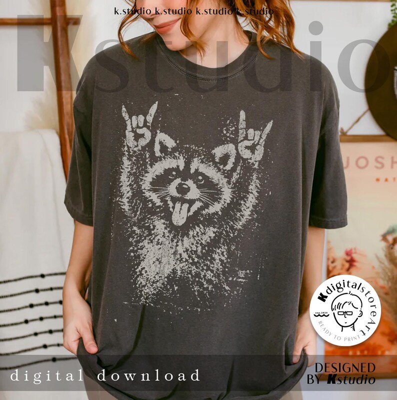 Pu&ograve; includere: T-shirt grigio scuro con una grafica usurata di un procione che fa gesti rock and roll. Il procione ha la lingua di fuori. Le parole "digital download" sono in basso. Design di Kstudio.