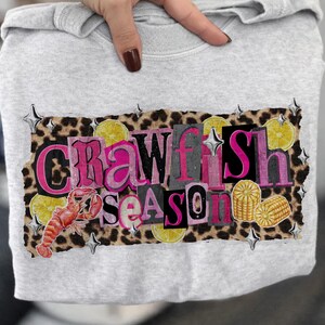 May include: A light gray sweatshirt featuring a graphic that reads "Crawfish Season" in pink and gray letters. The design includes a crawfish, lemons, and corn on a leopard print background.
