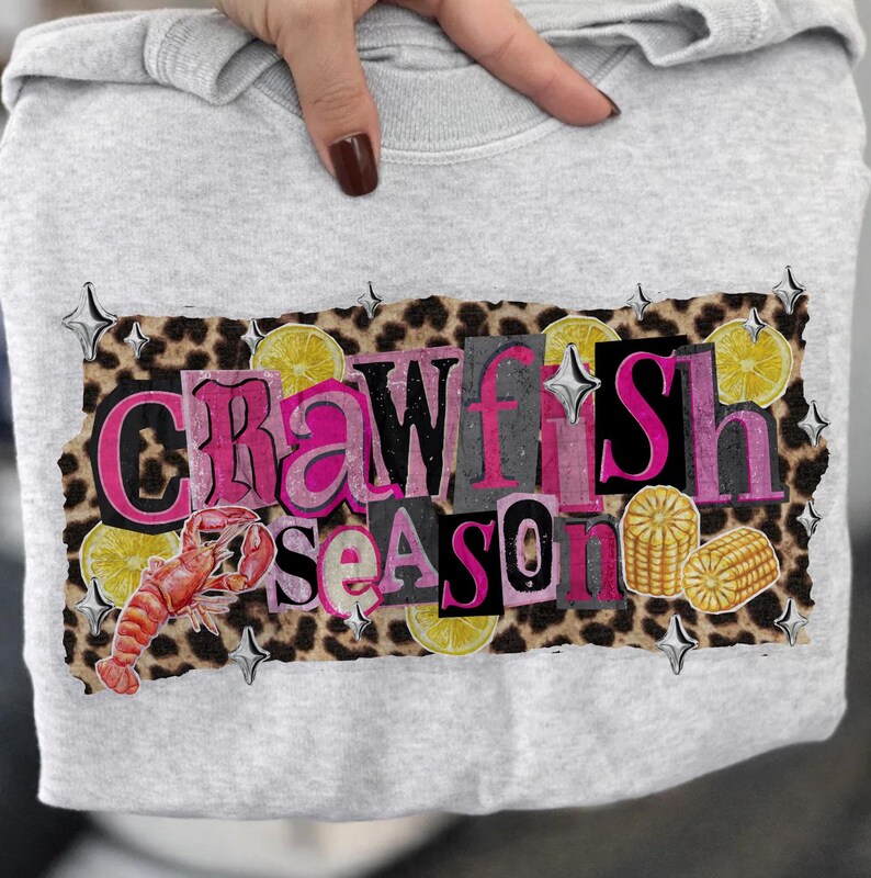 May include: A light gray sweatshirt featuring a graphic that reads "Crawfish Season" in pink and gray letters. The design includes a crawfish, lemons, and corn on a leopard print background.