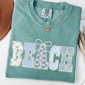 May include: A teal Comfort Colors t-shirt with the word "BEACH" in a decorative font. The letters are filled with patterns of seashells, starfish, and floral designs in shades of blue and white, outlined with gold beads. A gold necklace with a pendant rests on the shirt.