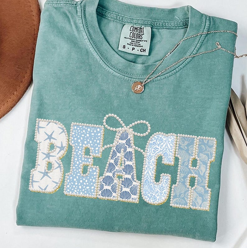 May include: A teal Comfort Colors t-shirt with the word "BEACH" in a decorative font. The letters are filled with patterns of seashells, starfish, and floral designs in shades of blue and white, outlined with gold beads. A gold necklace with a pendant rests on the shirt.