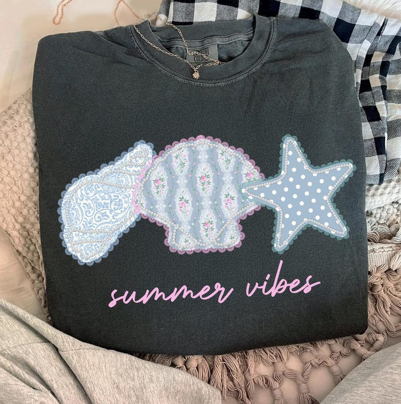 May include: Dark gray sweatshirt with a seashell and star design. The seashell and star are light blue with floral and polka dot patterns. The text "summer vibes" is in pink script below the design.