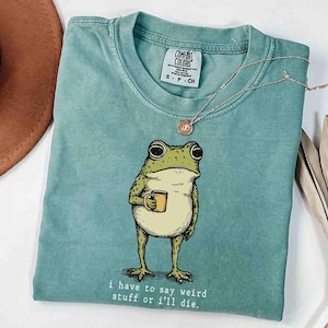 May include: A seafoam green t-shirt featuring a cartoon frog holding a yellow mug. The frog has large eyes and is standing upright. The text "i have to say weird stuff or i'll die" is printed below the frog. A silver necklace is draped across the shirt.