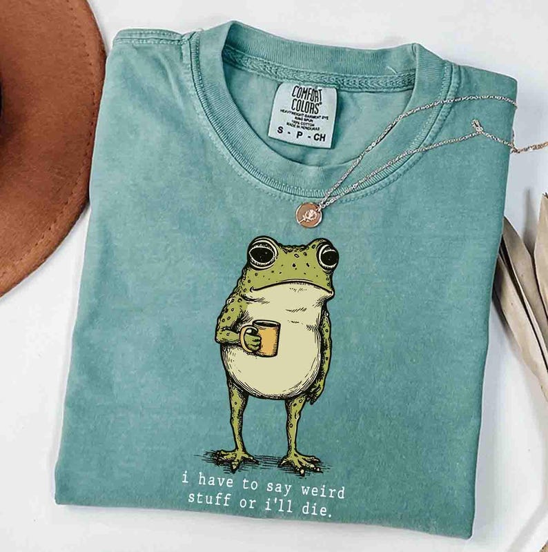 May include: A seafoam green t-shirt featuring a cartoon frog holding a yellow mug. The frog has large eyes and is standing upright. The text "i have to say weird stuff or i'll die" is printed below the frog. A silver necklace is draped across the shirt.