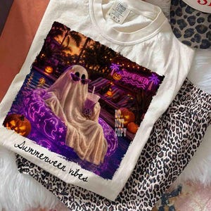 May include: Cream-colored t-shirt with a graphic featuring a ghost in sunglasses relaxing in a pool float, holding a drink. The image has a neon sign that says "Summerween". The words "Summerween vibes" are printed on the shirt.
