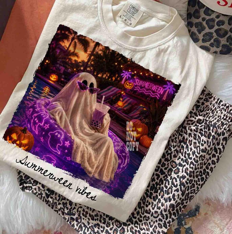 May include: Cream-colored t-shirt with a graphic featuring a ghost in sunglasses relaxing in a pool float, holding a drink. The image has a neon sign that says "Summerween". The words "Summerween vibes" are printed on the shirt.