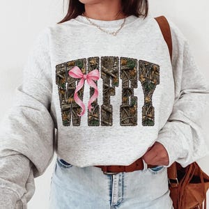Puede incluir: Sudadera gris claro con la palabra "WIFEY" en un patr&oacute;n de camuflaje y un lazo rosa. La sudadera tiene mangas largas y cuello redondo.