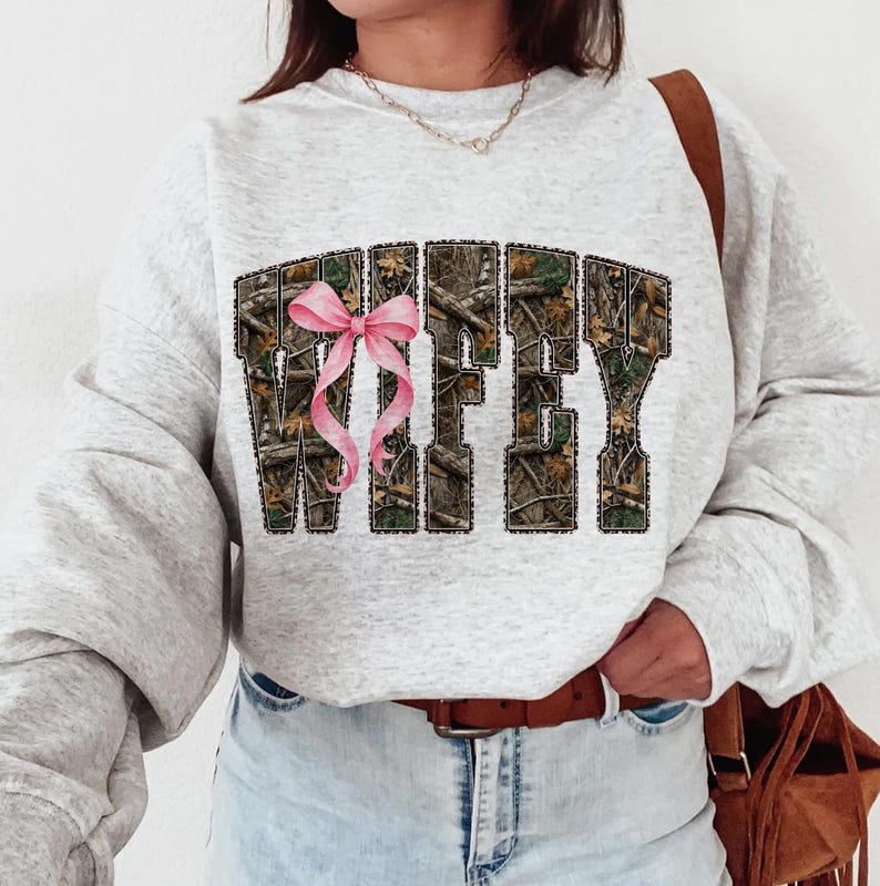 Puede incluir: Sudadera gris claro con la palabra "WIFEY" en un patr&oacute;n de camuflaje y un lazo rosa. La sudadera tiene mangas largas y cuello redondo.