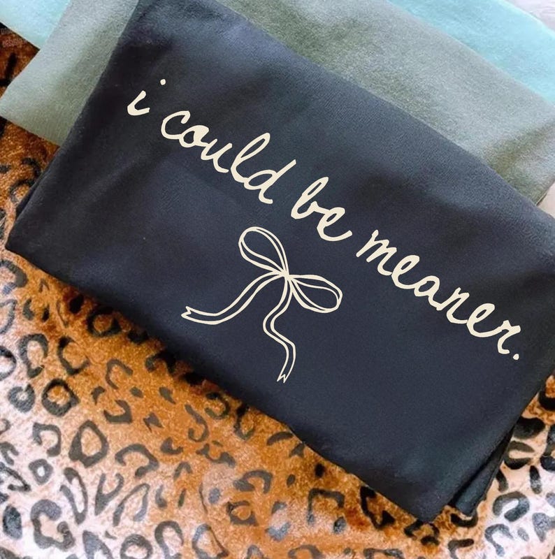 May include: Black tote bag with the phrase "i could be meaner" in a handwritten style, along with a bow graphic. The bag is on a leopard print surface, with a few other fabric colors visible in the background.