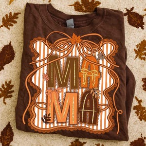 Op de afbeelding: Bruin T-shirt met het woord "MAMA" in decoratieve letters. Het ontwerp gebruikt herfstkleuren en -elementen. Het shirt is opgevouwen, een label is zichtbaar.