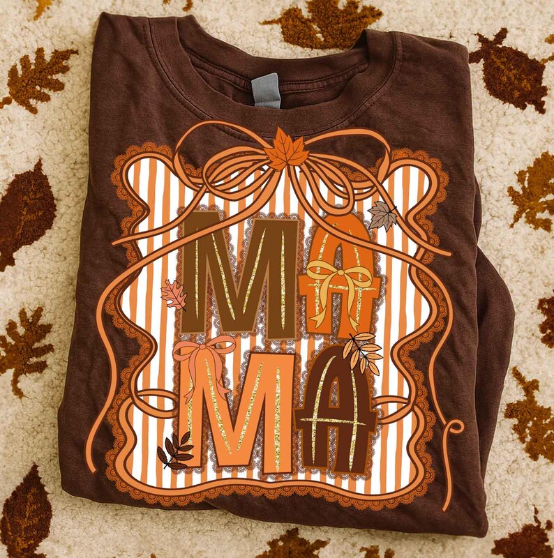 Op de afbeelding: Bruin T-shirt met het woord "MAMA" in decoratieve letters. Het ontwerp gebruikt herfstkleuren en -elementen. Het shirt is opgevouwen, een label is zichtbaar.