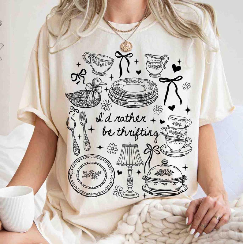 May include: Cream-colored t-shirt featuring a black and white illustration of teacups, plates, a duck, and the phrase "I'd rather be thrifting." The design includes floral and bow accents, creating a vintage aesthetic.
