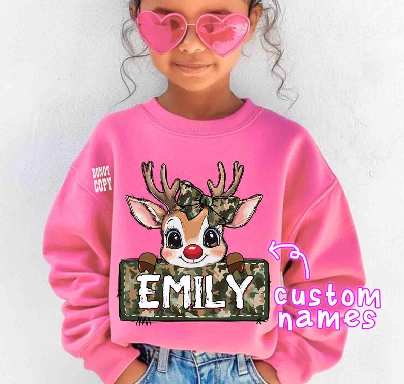 May include: Pink sweatshirt featuring a cartoon reindeer with a camouflage bow and a sign that says "EMILY". The sweatshirt has the text "DO NOT COPY" on the upper left side. The words "custom names" are also visible.