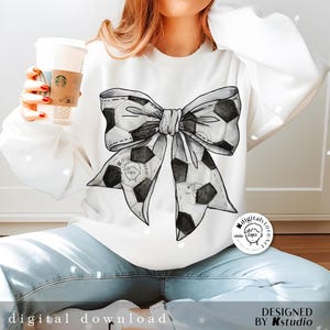 May include: A white sweatshirt with a black and white watercolor soccer ball bow design. The sweatshirt is being worn by a person. The design includes the text "Digitalstore" in a circle.