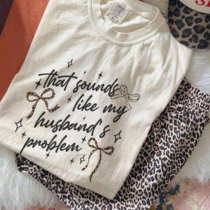 May include: Cream-colored t-shirt with the phrase "That sounds like my husband's problem" in black script, accented with leopard print bows and stars. Paired with leopard print pajama pants. The shirt has a Comfort Colors tag.