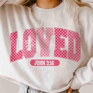 May include: A light gray sweatshirt with the word "LOVED" in large, pink, checkered letters. Below the word is the text "JOHN 3:16" in a pink rectangular box. The sweatshirt is a casual style.