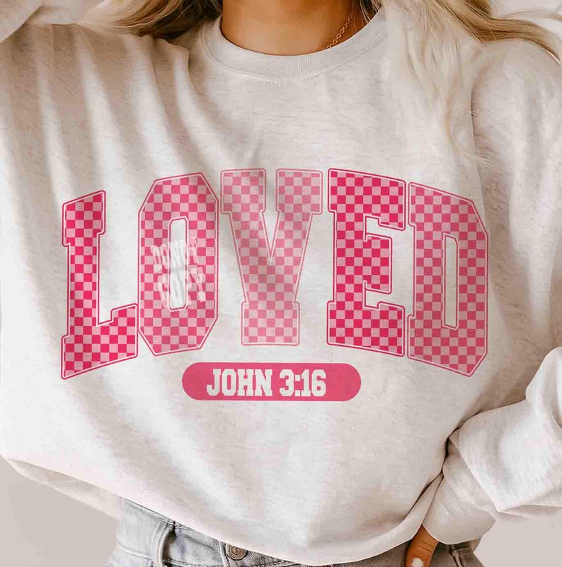 May include: A light gray sweatshirt with the word "LOVED" in large, pink, checkered letters. Below the word is the text "JOHN 3:16" in a pink rectangular box. The sweatshirt is a casual style.