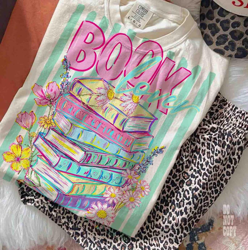 May include: Cream-colored t-shirt with the words "BOOK LOVER" in pink and blue, stacked books, and floral accents. The shirt is paired with leopard print pants. The shirt has a Comfort Colors tag.