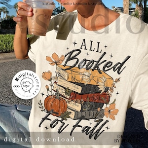 Puede incluir: Camiseta color crema con un gráfico temático de otoño. El diseño presenta una pila de libros, una calabaza y detalles florales, con el texto "ALL Booked For Fall". La camiseta incluye un logotipo de descarga digital.