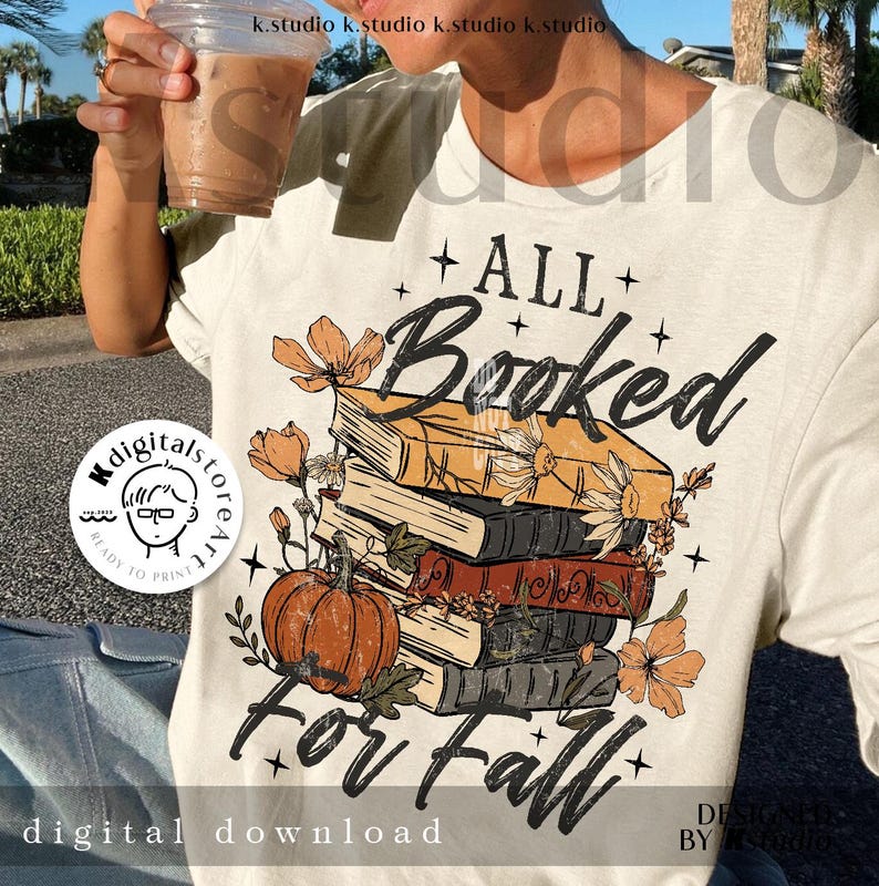 May include: Cream-colored t-shirt with a fall-themed graphic. The design features a stack of books, a pumpkin, and floral accents, with the text "ALL Booked For Fall." The shirt includes a digital download logo.