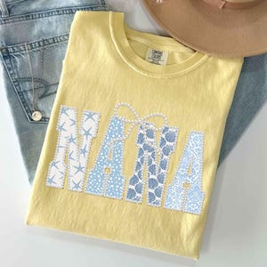 Puede incluir: Una camiseta amarilla Comfort Colors con la palabra "NANA" en letras estampadas blancas y azules. La "N" est&aacute; decorada con estrellas de mar blancas, la "A" con lunares azules y la otra "A" con conchas azules. Tambi&eacute;n se ven un collar dorado y un sombrero beige.