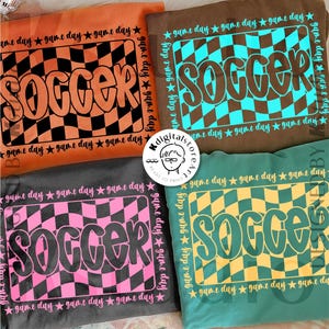 Op de afbeelding: Vier voetbal-thema sweatshirts in oranje, bruin, grijs en blauwgroen. Elk shirt heeft het woord "SOCCER" in een bubbellettertype over een geruit patroon. De zin "game day" en sterren omringen het ontwerp.